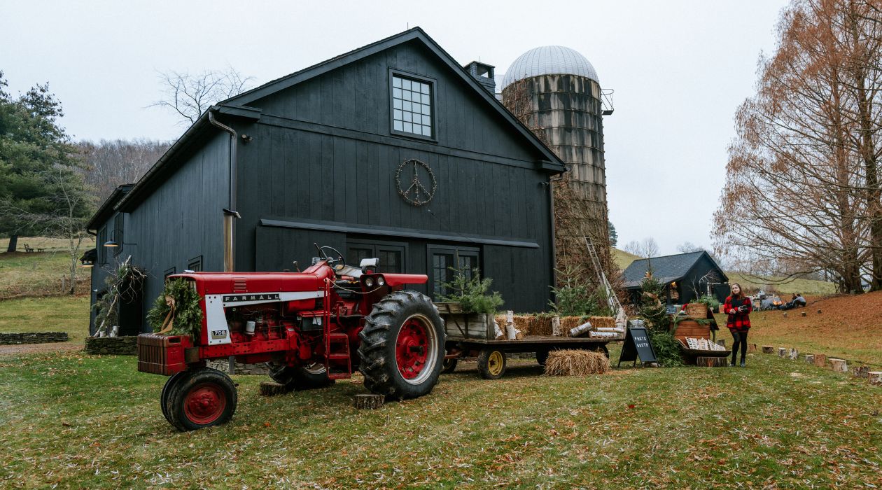 The Black Barn Winter Market at SkyRise Farm in Millerton, with a red and white Farmall tractor and a trailer carrying hay bales and Christmas trees parked in front of the black barn and white silo. Photo by Peter Crosby